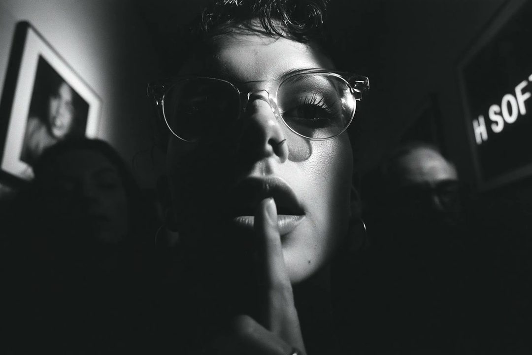 A black and white close-up portrait of a person wearing glasses, with a finger on their lips, in dramatic lighting.