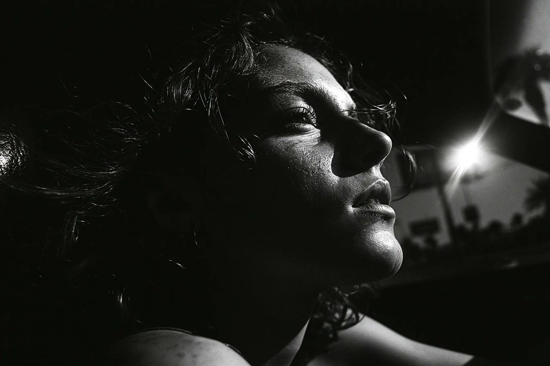 A black and white close-up portrait of a person with curly hair, looking upwards with dramatic lighting.