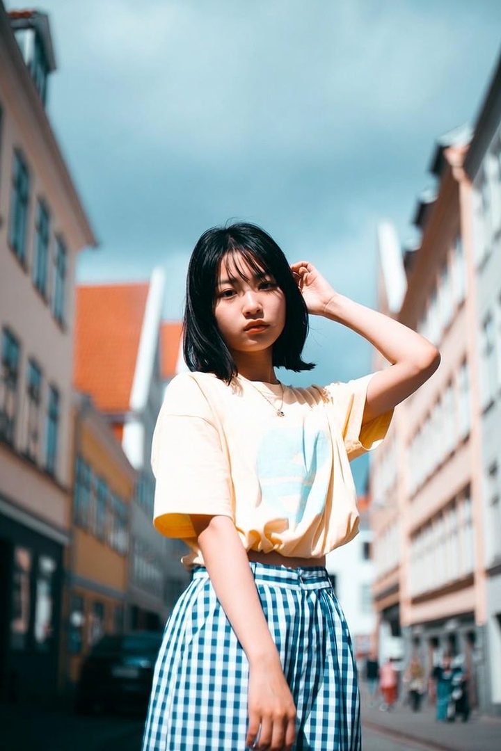 A young woman with short black hair and bangs, wearing a yellow t-shirt and a blue and white checkered skirt, stands on a street with buildings in the background.