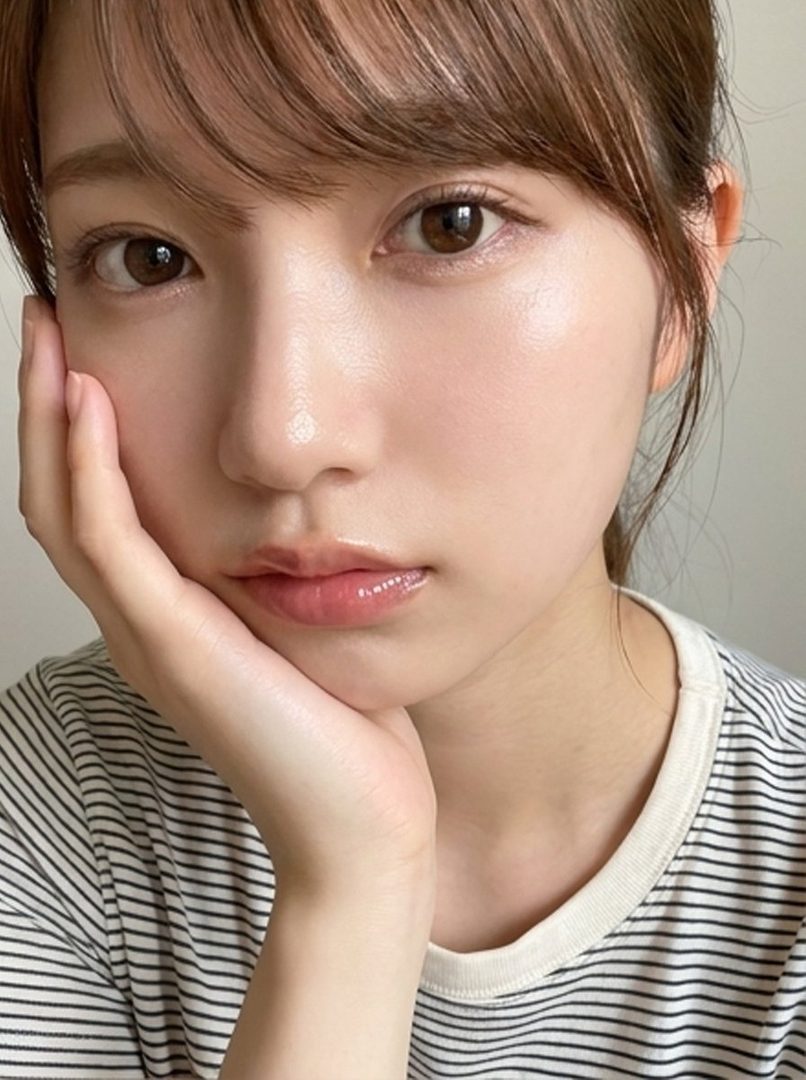 A close-up portrait of a young woman with brown hair and bangs, wearing a striped shirt, resting her chin on her hand.