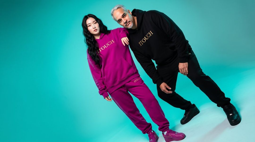 A man and a woman pose in a studio wearing matching hoodies and sweatpants, with the woman in purple and the man in black.
