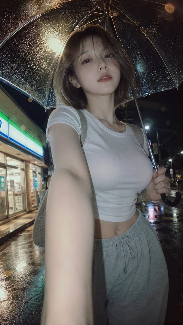 A young woman takes a selfie at night on a rainy street, holding an umbrella and wearing a white crop top and grey sweatpants.