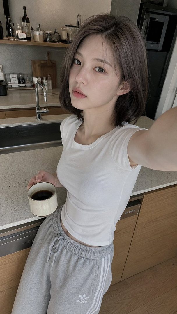 A young woman with short brown hair takes a selfie in a kitchen, holding a cup of coffee.