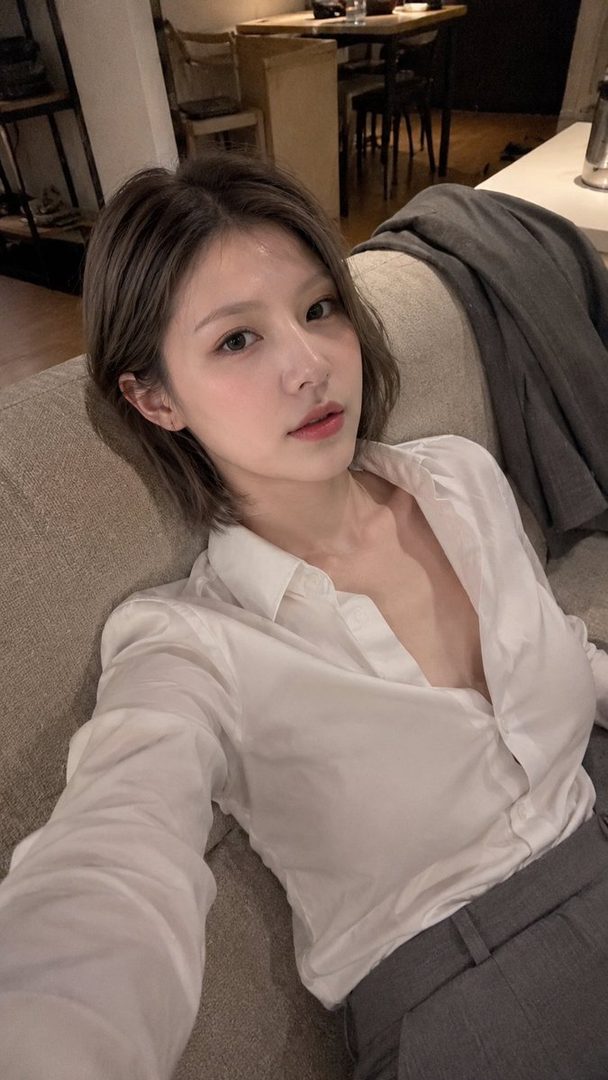 A photorealistic selfie of an East Asian woman with short brown hair, wearing a white blouse and grey trousers, reclining on a sofa indoors.