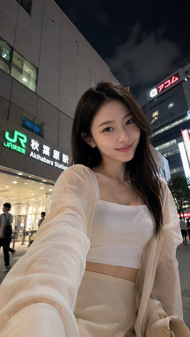 A young woman takes a selfie at night in front of Akihabara Station in Tokyo, wearing a white crop top and a sheer light-colored jacket.