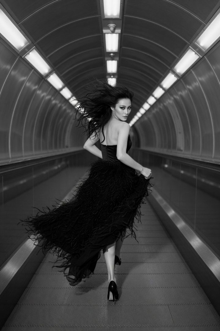 A photorealistic black and white image of an East Asian woman in a black feathered dress running through a futuristic tunnel, looking back at the camera.