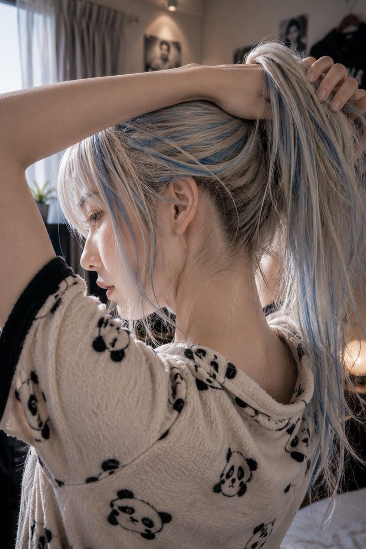 A woman with blonde and blue hair ties her hair into a ponytail, wearing a panda-patterned top indoors.