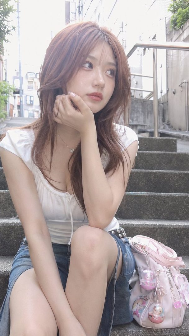 A young woman with brown hair sits on stone steps outdoors, wearing a white top and denim skirt, with a pink backpack beside her.