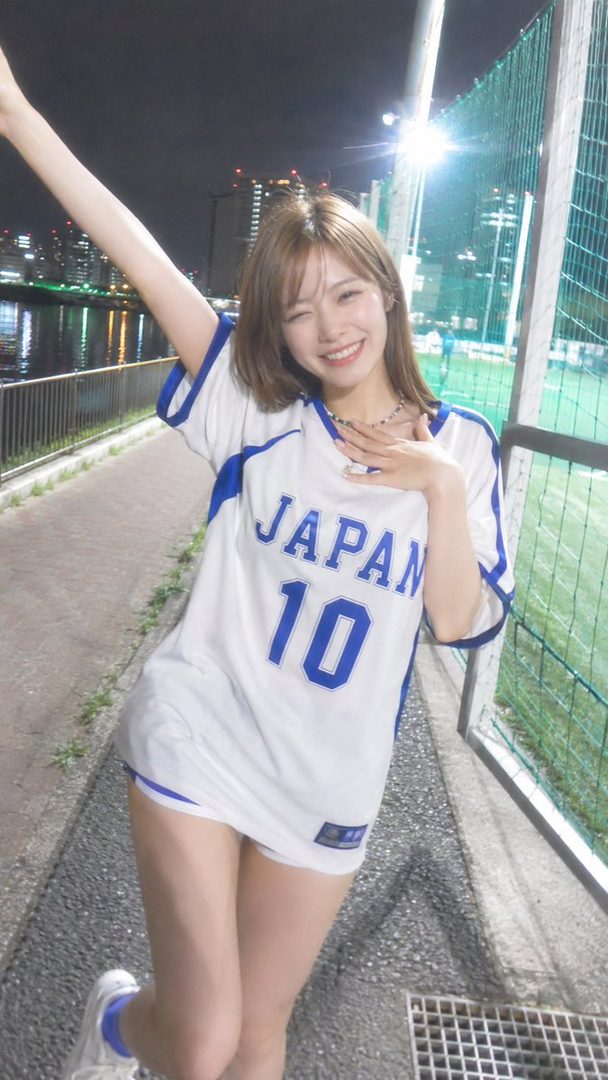 A young woman in a Japan jersey with number 10 smiles playfully at night by a river and a sports field.