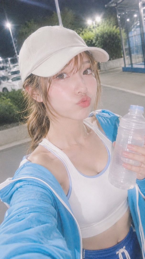 A young woman in sportswear and a cap takes a selfie outdoors at night, holding a water bottle and making a kissy face.