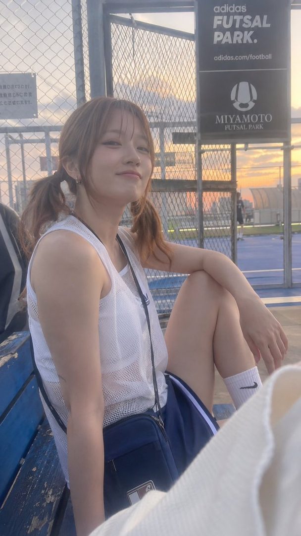A young woman with brown hair in pigtails and bangs, wearing sportswear, sits outdoors at a futsal park during golden hour.