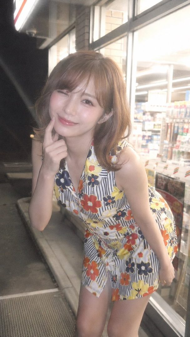 A young woman with brown hair winks at the camera while standing outside a convenience store at night, wearing a floral and striped dress.