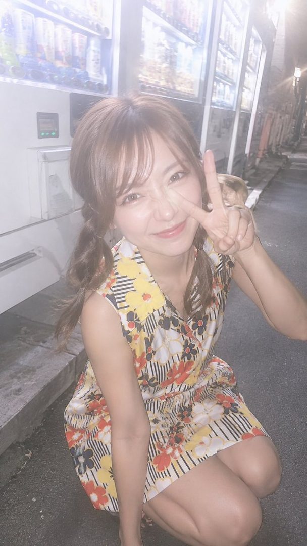 A young woman with brown twintails and bangs, wearing a floral dress, crouches on a street at night, making a peace sign.