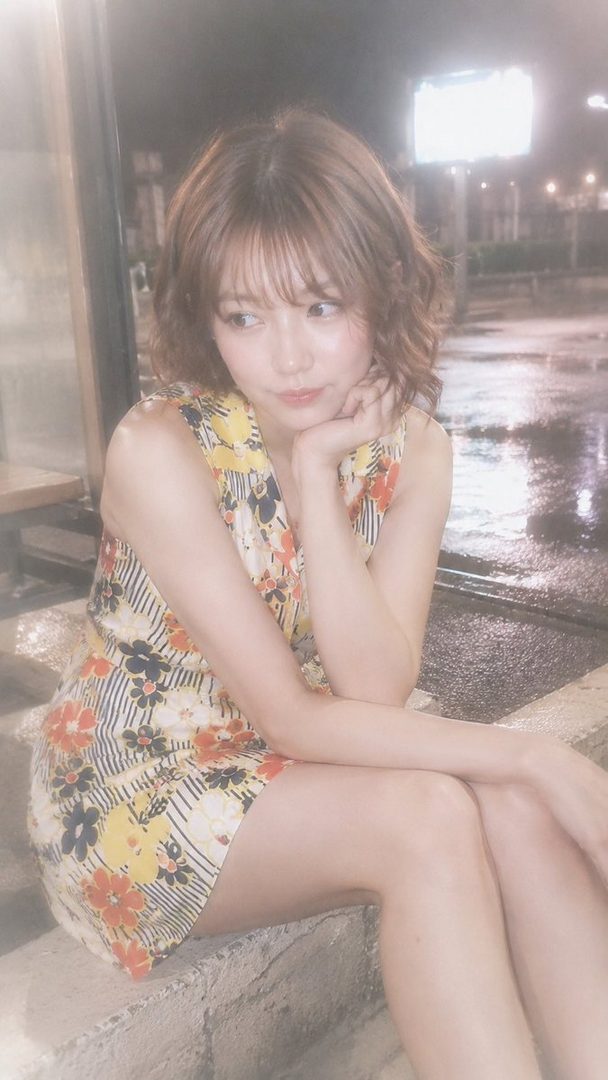 A young woman with short curly brown hair and bangs, wearing a floral dress, sits on a stone ledge at night with a dreamy expression.