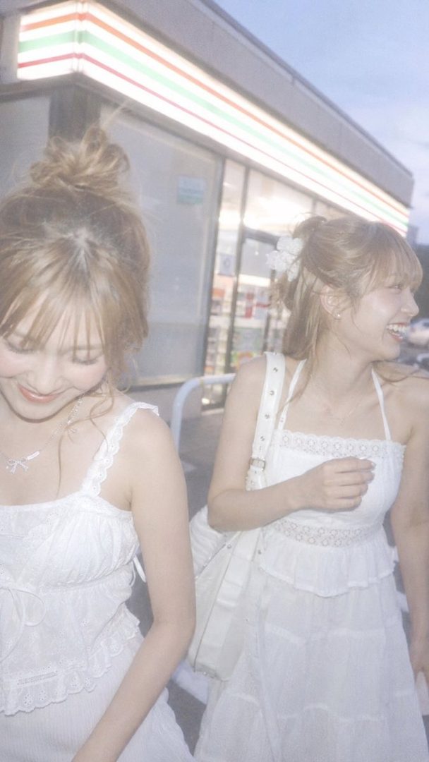 Two young women in white dresses are laughing and walking at night in front of a convenience store.