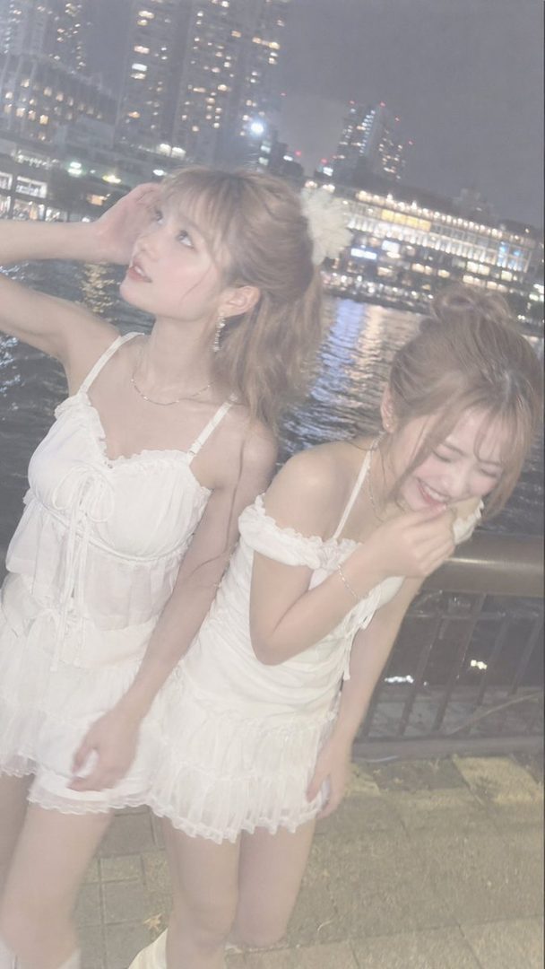 Two young women in white dresses pose by a city waterfront at night, with city lights in the background.