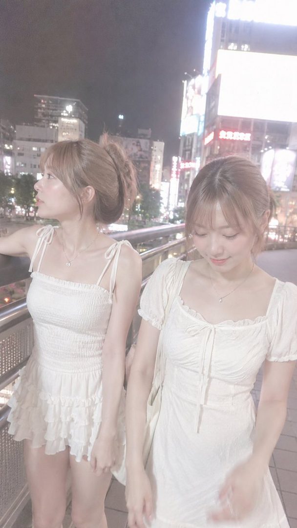 Two young women in white dresses are standing on a bridge in a city at night, with one looking into the distance and the other looking down.