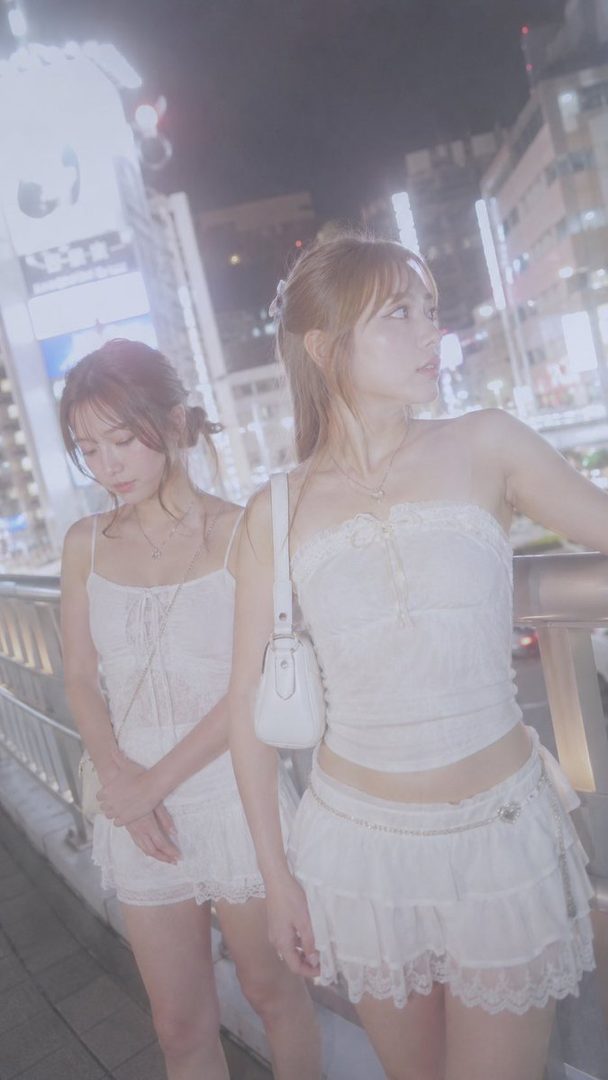 Two young women in white lace outfits stand on a city street at night, with soft lighting and a dreamy atmosphere.