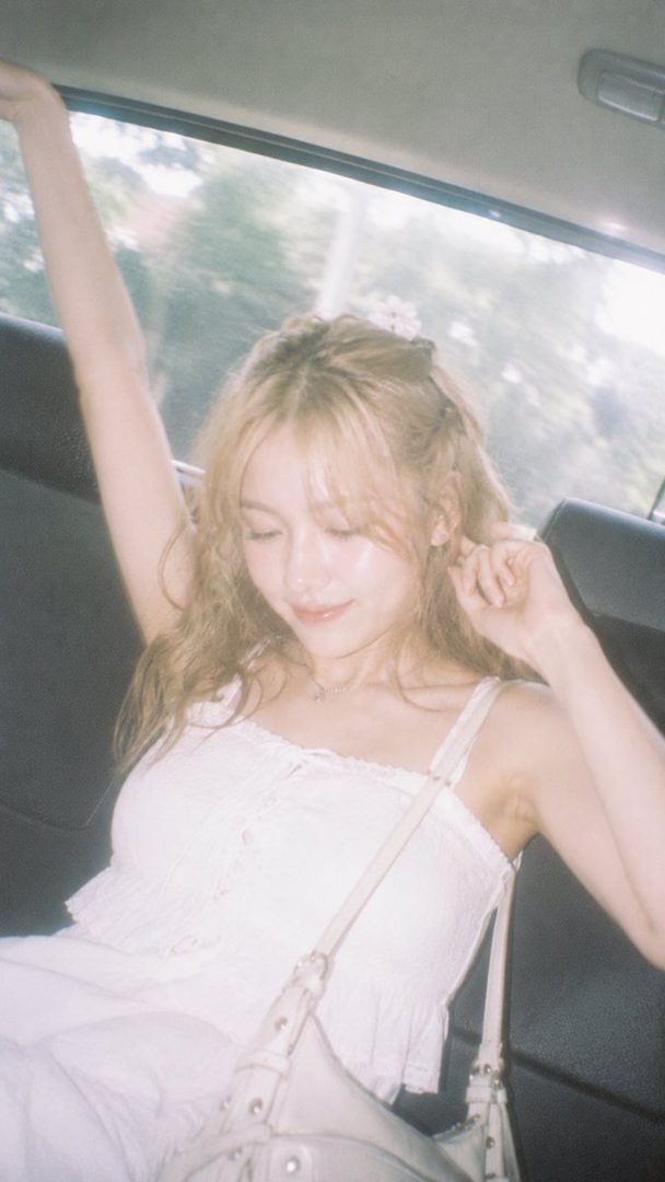A young woman with blonde hair and a white top is sitting in a car, looking down with a dreamy expression.