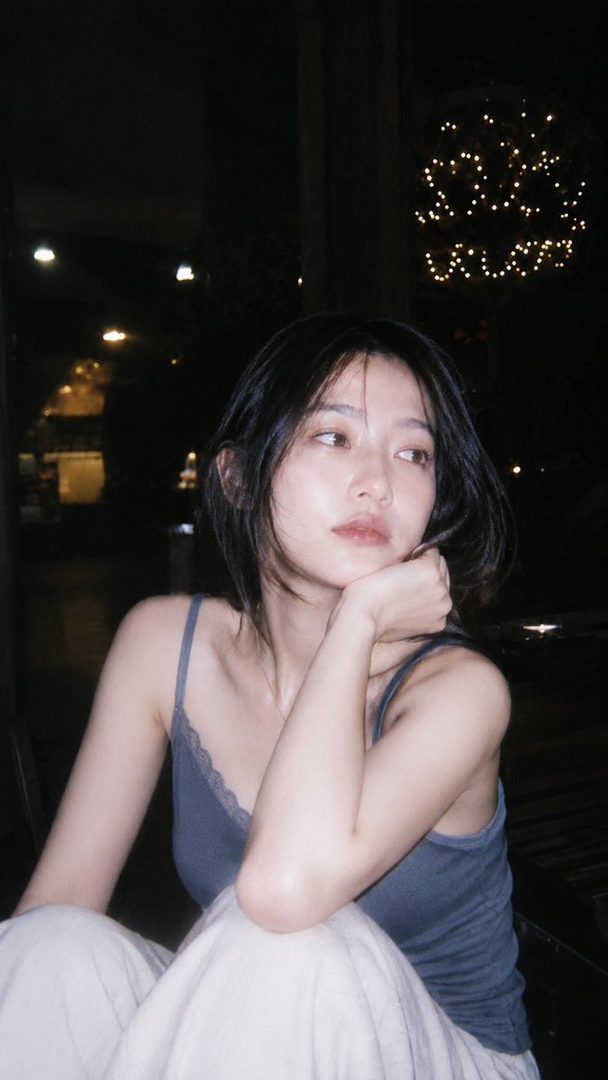 A photorealistic portrait of a young East Asian woman with short black hair, wearing a casual top, looking thoughtfully into the distance at night.