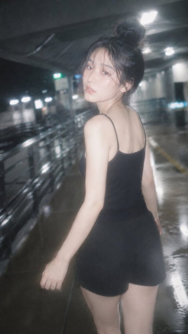 A young woman with black hair tied in a bun looks back over her shoulder in a dimly lit outdoor setting.