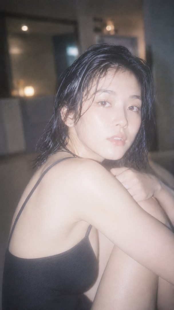 A woman with wet, short black hair and a black tank top looks directly at the camera in a soft-lit indoor setting.