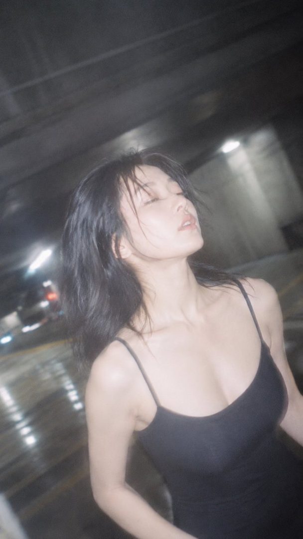 A woman with black hair and a black tank top stands with her eyes closed in a dimly lit indoor setting.