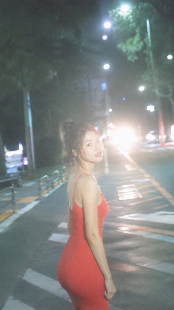 A young woman in a red dress stands on a city street at night, looking back at the camera with car lights in the background.