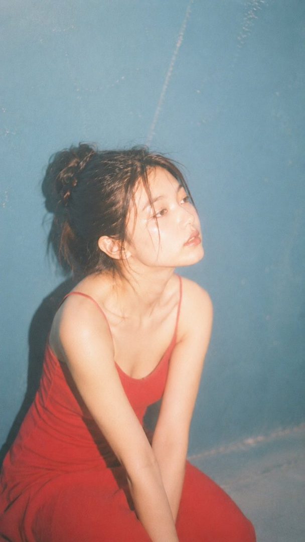 A young East Asian woman in a red dress with her hair tied up, looking upwards against a blue background.