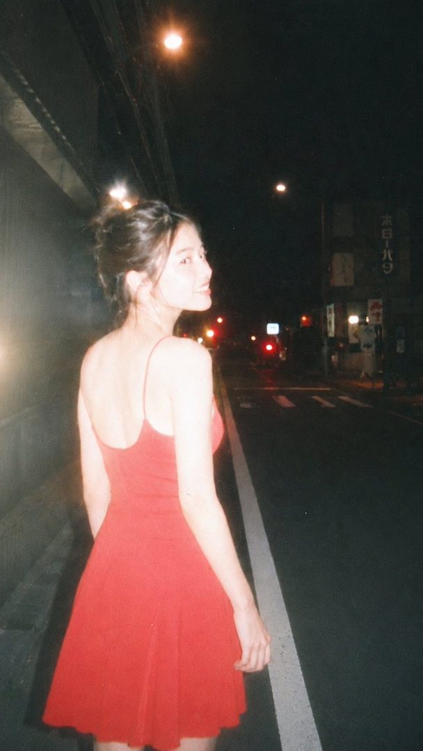 A young woman in a red dress looks back over her shoulder on a dimly lit street at night.