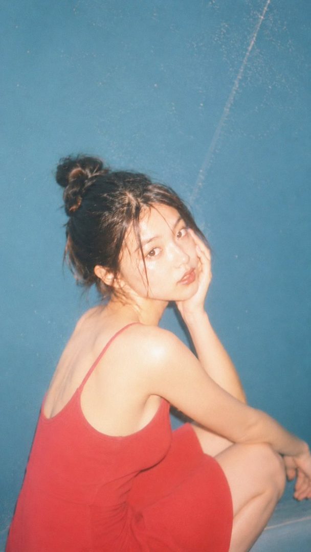 A young woman with dark hair tied up, wearing a red dress, looks over her shoulder with a dreamy expression against a blue background.