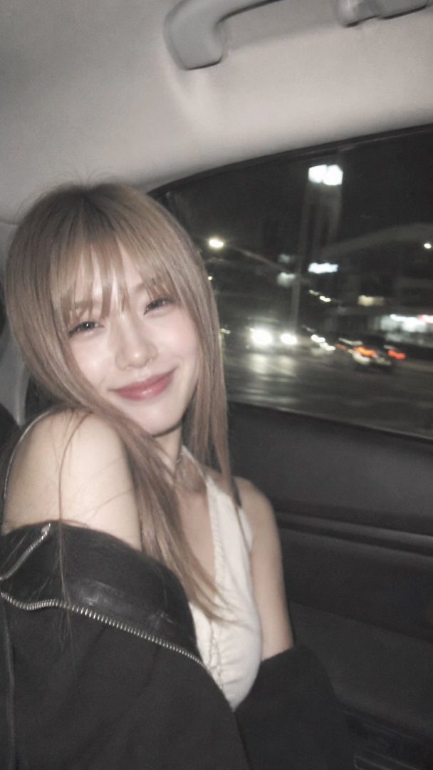 A smiling woman with blonde hair and bangs sits in a car at night, with city lights visible outside the window.