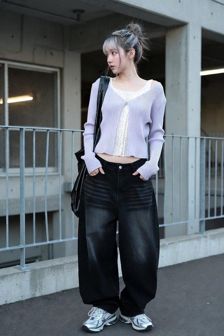 A young woman with silver hair tied in a bun, wearing a light purple knit cardigan over a white lace top and baggy black jeans, stands outdoors.