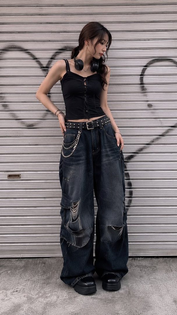 A young woman with long brown hair and headphones around her neck poses in a black tank top and ripped baggy jeans against a metal shutter with graffiti.