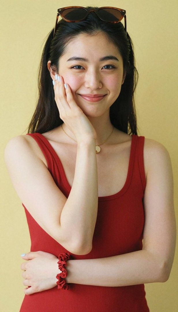 A young woman with long black hair and sunglasses on her head smiles while wearing a red tank top against a yellow background.