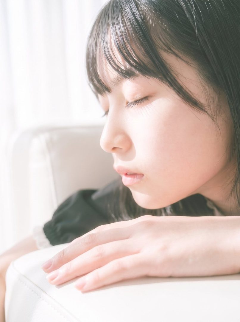 A serene portrait of a young woman with black hair and bangs, eyes closed, bathed in soft light indoors.