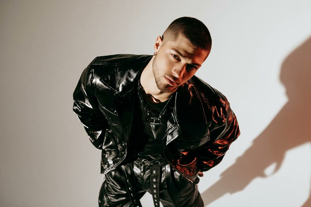 A man with short brown hair and a beard wearing a black shiny jacket and pants, looking up at the camera in a studio.