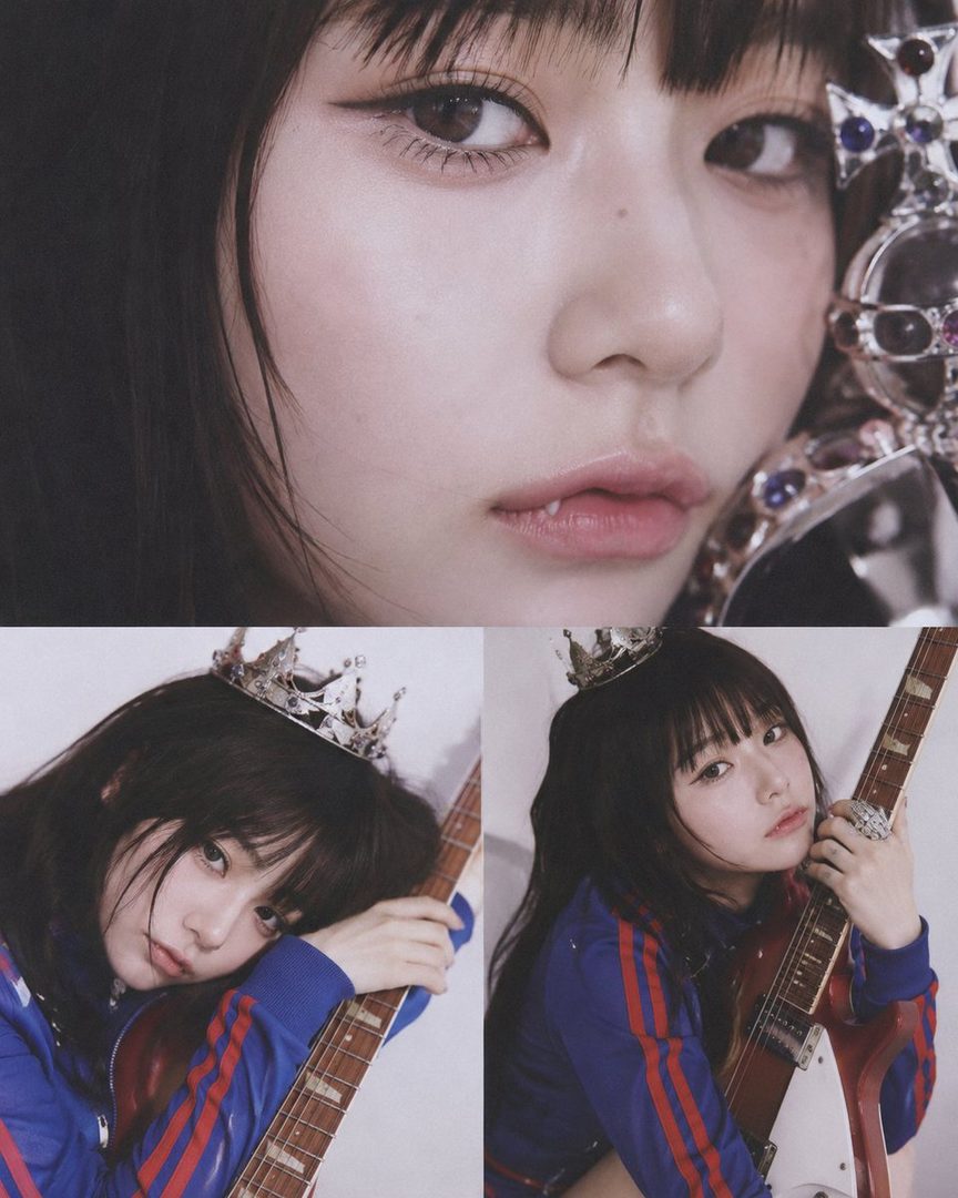 A close-up portrait of a woman with black hair and bangs, wearing a blue and red tracksuit, holding an electric guitar and a small crown on her head.