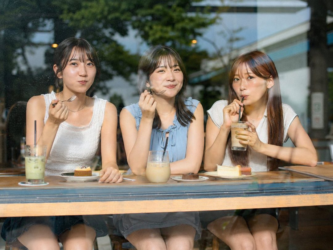 Three young women are sitting at a table in a cafe, eating cake and drinking beverages, looking out through a window.
