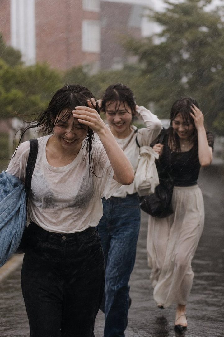 Three young women are laughing and running in the rain on a street, with their clothes and hair wet.