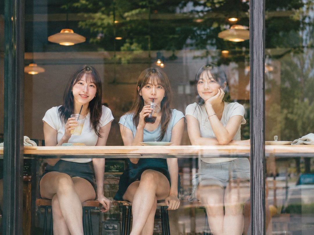 Three young women are sitting at a counter inside a cafe, seen through a window, enjoying drinks on a sunny day.
