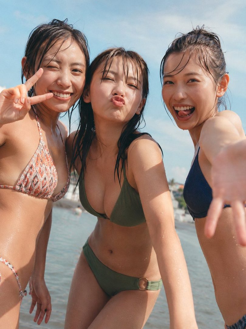 Three young women in swimsuits pose playfully for a selfie at the beach on a sunny day.