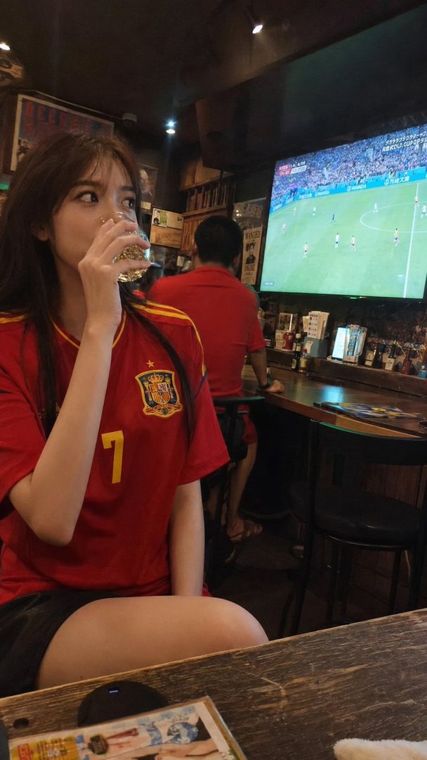 A young woman in a red Spain soccer jersey drinks from a glass while watching a World Cup match on a large screen in a dimly lit bar.