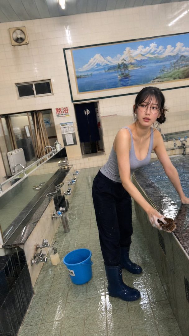 A young woman in glasses, a grey tank top, and dark pants is cleaning a bath area with a brush and soap, wearing blue rubber boots.