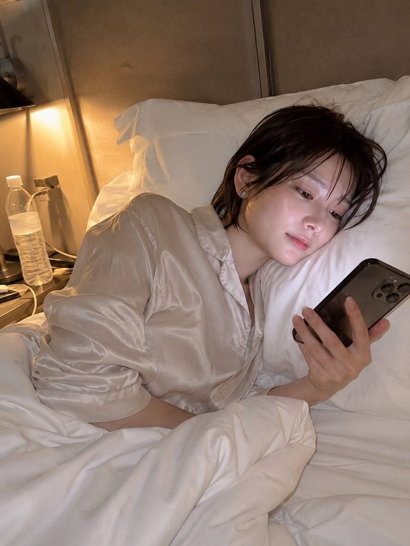 A woman with short brown hair is lying in bed, wearing light-colored pajamas and looking at her phone.