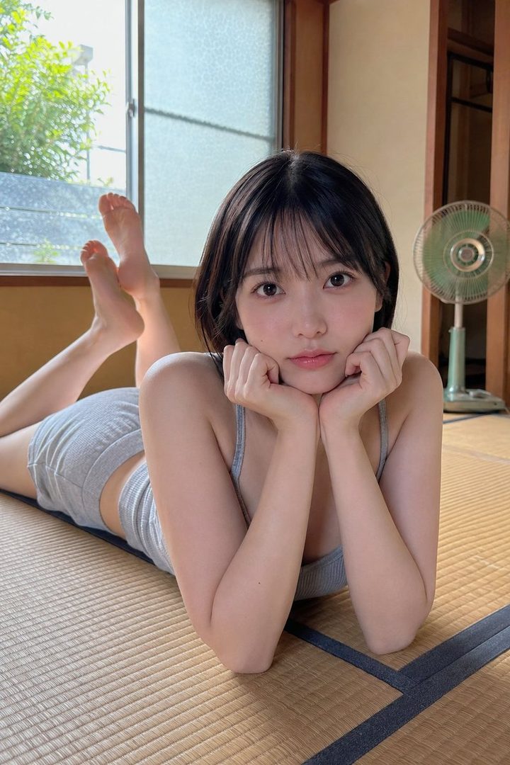 A young woman with short black hair lies on a tatami mat, wearing a grey tank top and shorts, looking directly at the camera.