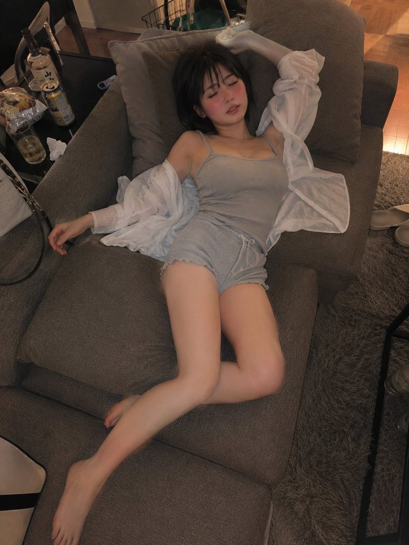 A young woman with short black hair is lying on a sofa, wearing a grey camisole and shorts, with a sheer white shirt draped over her.