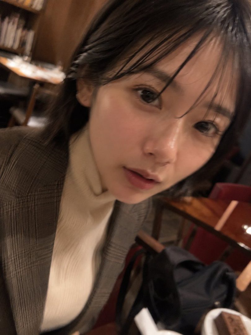 A photorealistic portrait of a young woman with dark hair and bangs, wearing a beige turtleneck and a plaid coat, looking directly at the camera in an indoor setting.