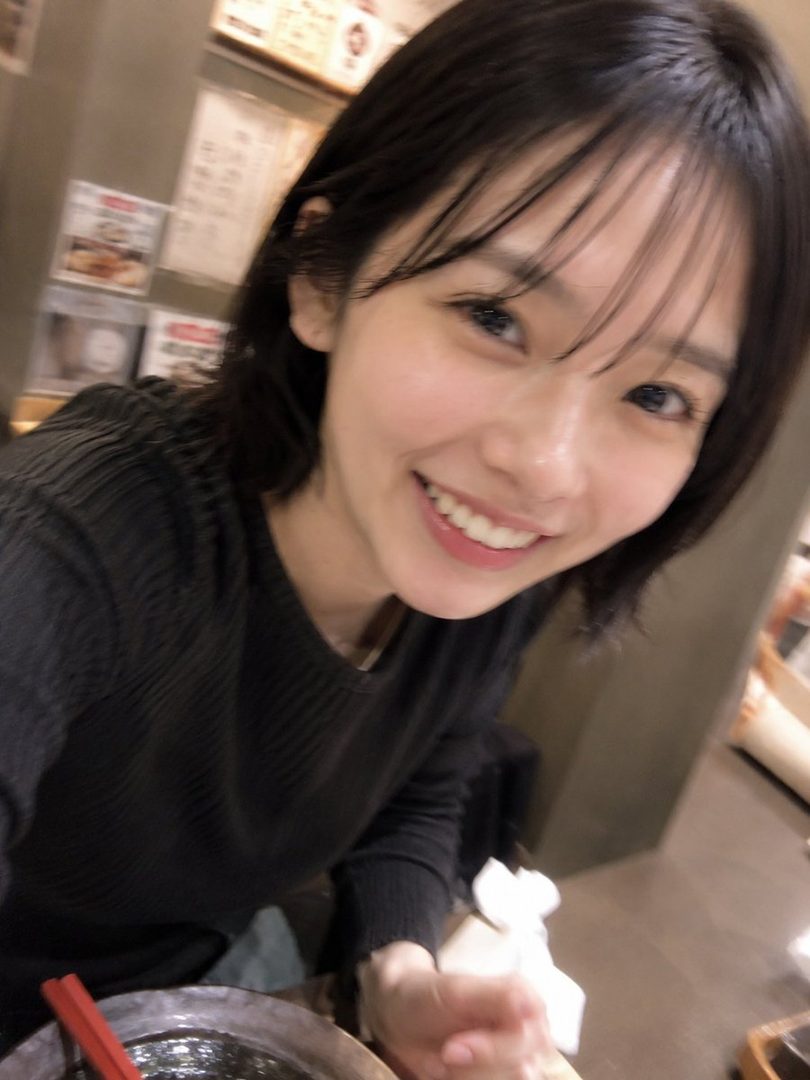 A smiling young woman with short black hair and bangs, wearing a black knit top, is seen indoors, possibly at a restaurant.
