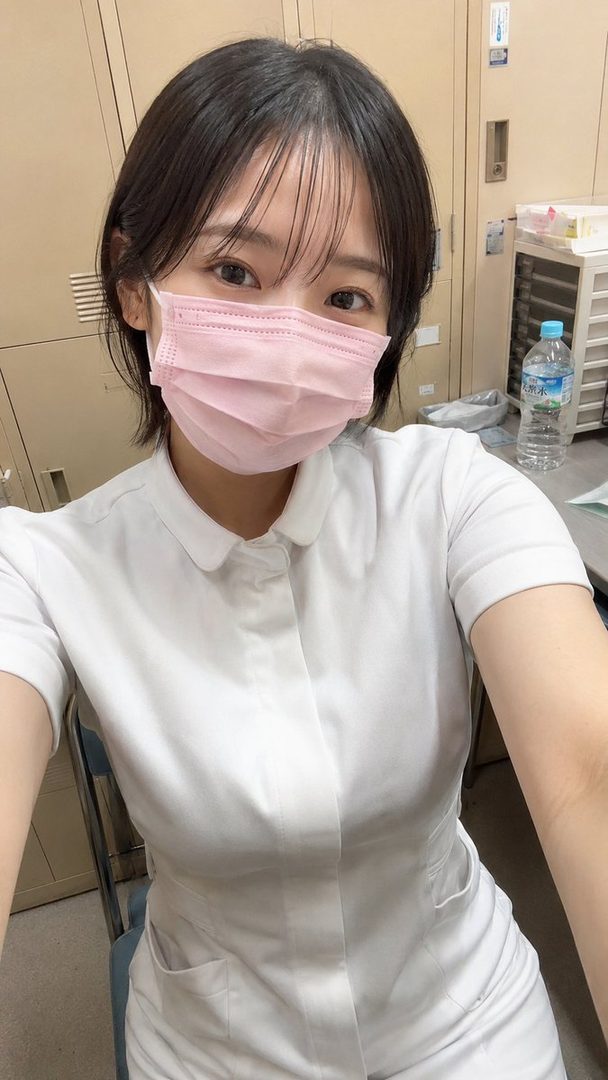 A young woman with short black hair and bangs, wearing a white nurse uniform and a pink face mask, takes a selfie indoors.